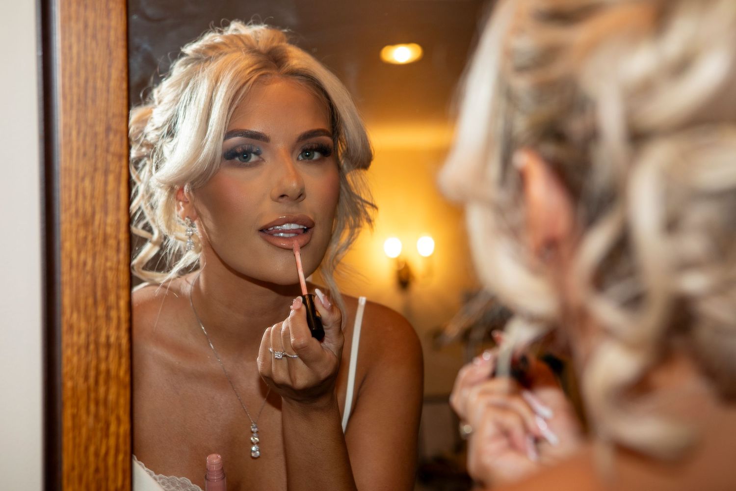 Blonde bride applying lip gloss while looking in a mirror during wedding day preparations.