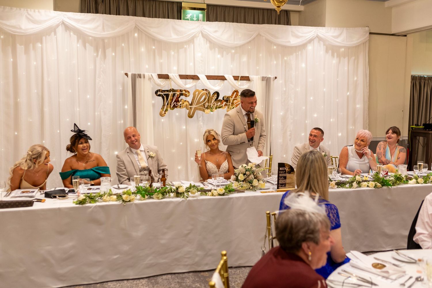 Groom giving a speech at wedding reception head table decorated with greenery and a personalized name sign backdrop.