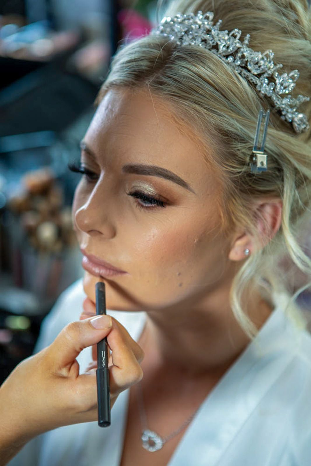 Bride getting lip liner applied by makeup artist, wearing crystal tiara and white robe for wedding day preparation.