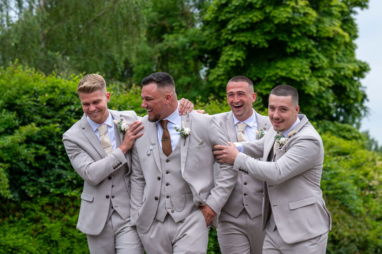 Four groomsmen in light grey suits laughing together outdoors with lush green hedges in the background at a wedding.