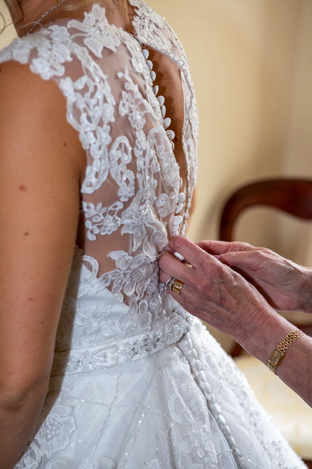 Elderly hands fastening the back of a bride's white lace wedding dress with intricate floral embroidery.