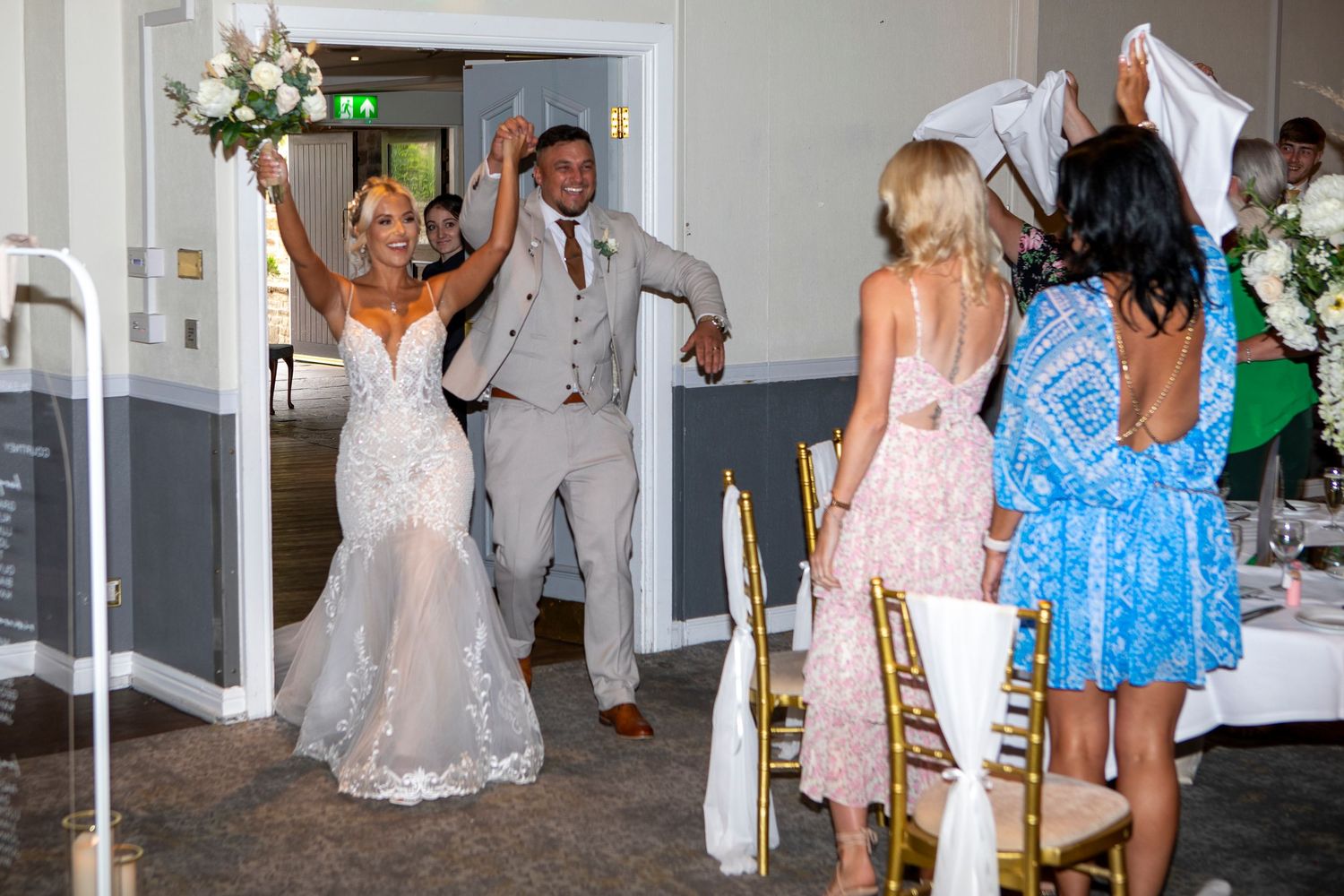 Bride and groom make grand entrance at wedding reception, guests cheering as bride raises bouquet in elegant venue.
