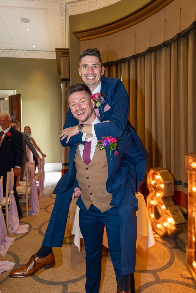 Two grooms in blue suits share a piggyback pose, smiling joyfully at their wedding reception.