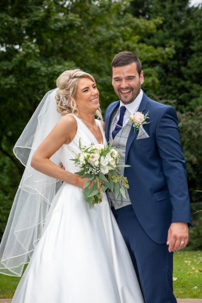 Happy bride and groom smile together outdoors, bride holding white floral bouquet with veil.