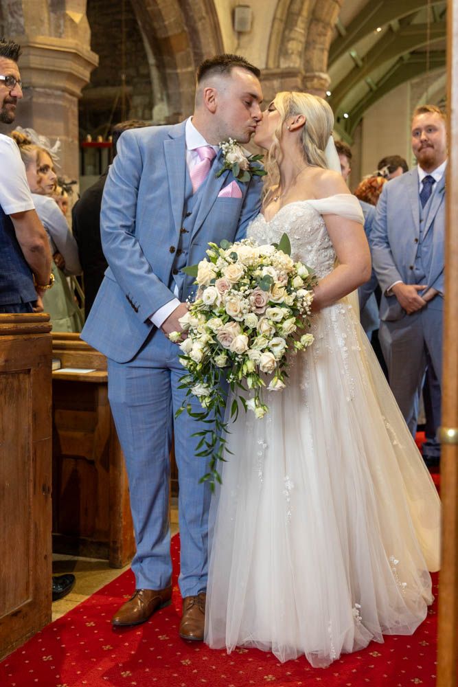 Bride and groom share first kiss at church altar, bride in off-shoulder gown holding pink bouquet, groom in blue suit.