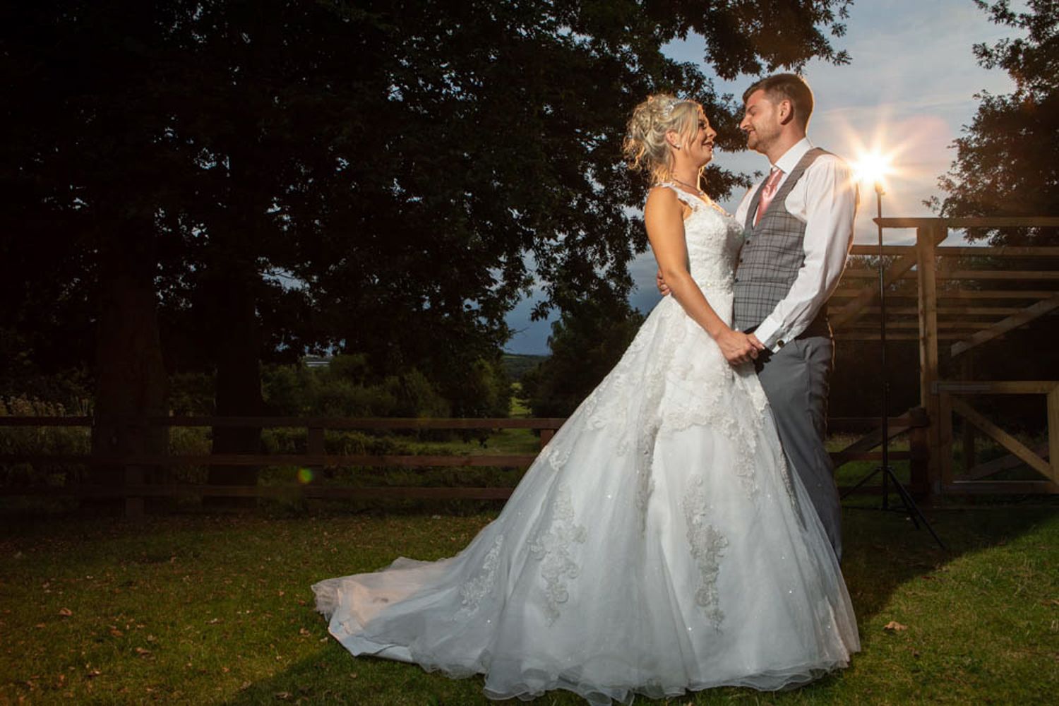 Bride and groom holding hands at sunset near a tree, bride wearing a white lace gown, groom in a gray vest.