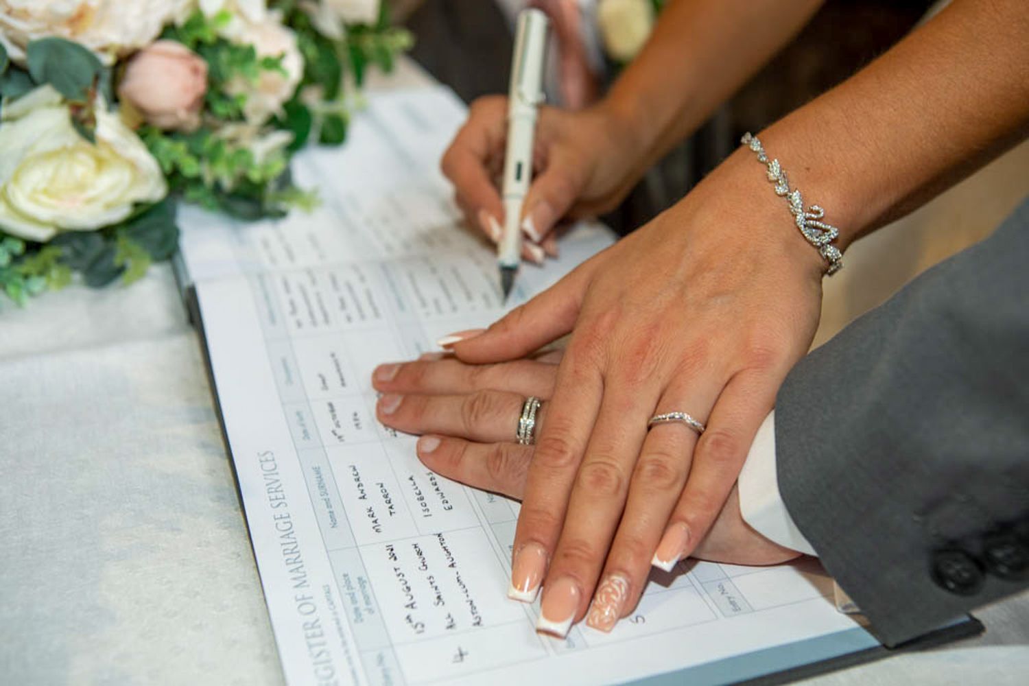 Newlyweds signing wedding certificate with diamond rings and bracelets visible, beside a white floral bouquet.