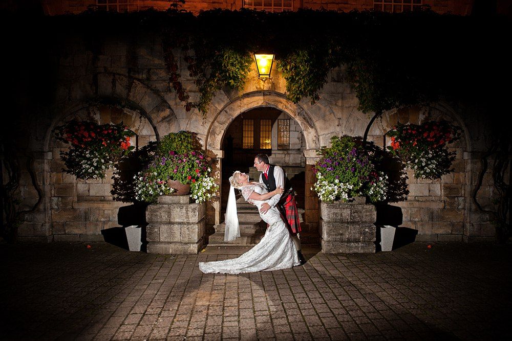Bride and groom share romantic kiss at night outside stone archway adorned with flowers and warm lighting.