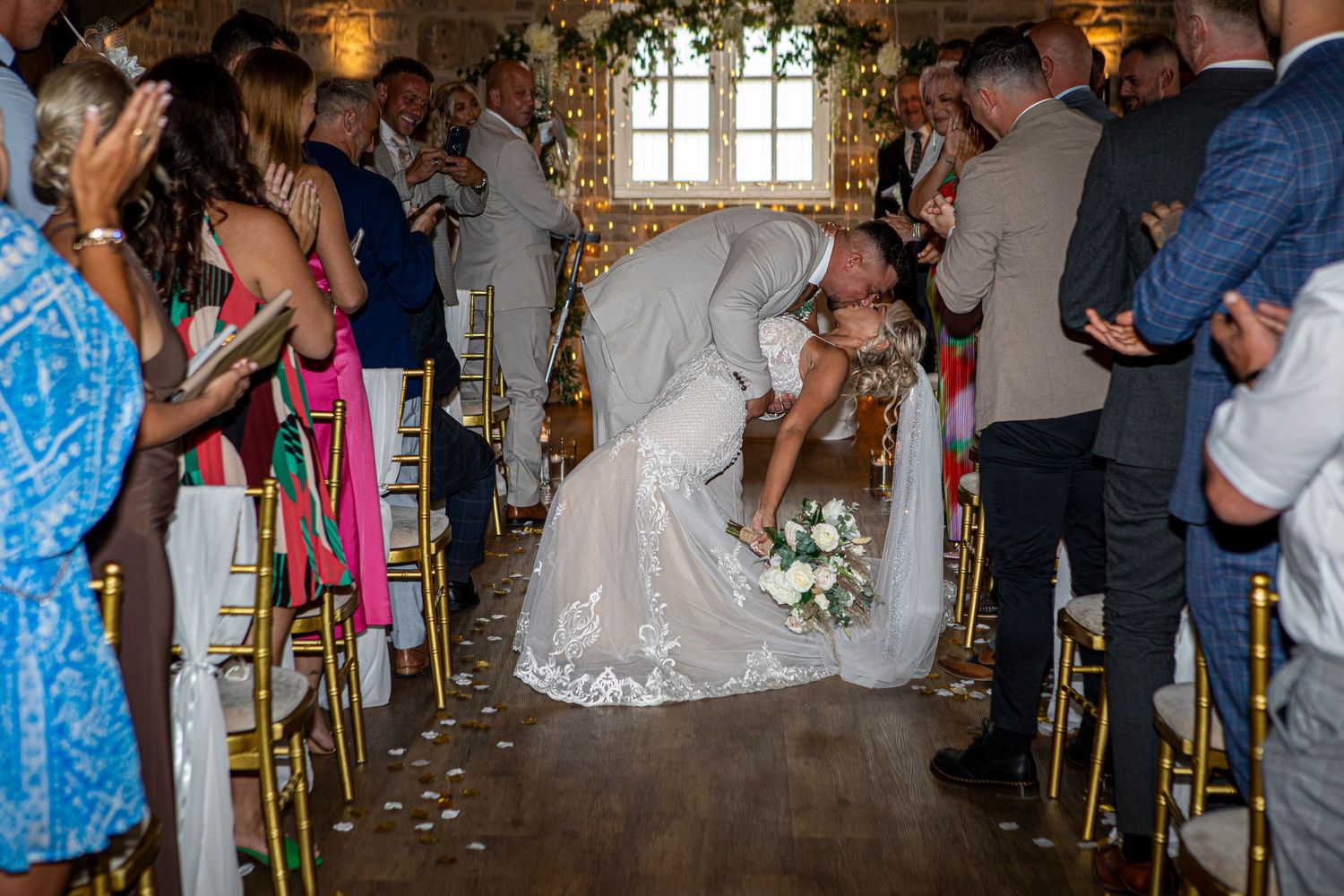 Bride and groom share a romantic dip kiss at wedding ceremony aisle surrounded by cheering guests in elegant venue.