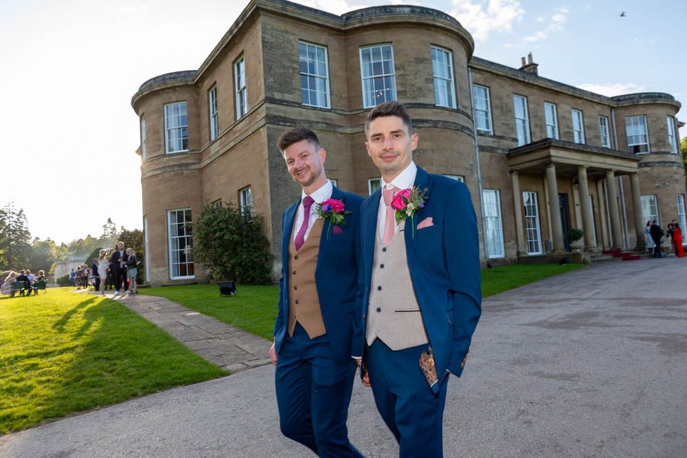 Two grooms in navy suits with pink boutonnieres pose outside an elegant stone manor house wedding venue.