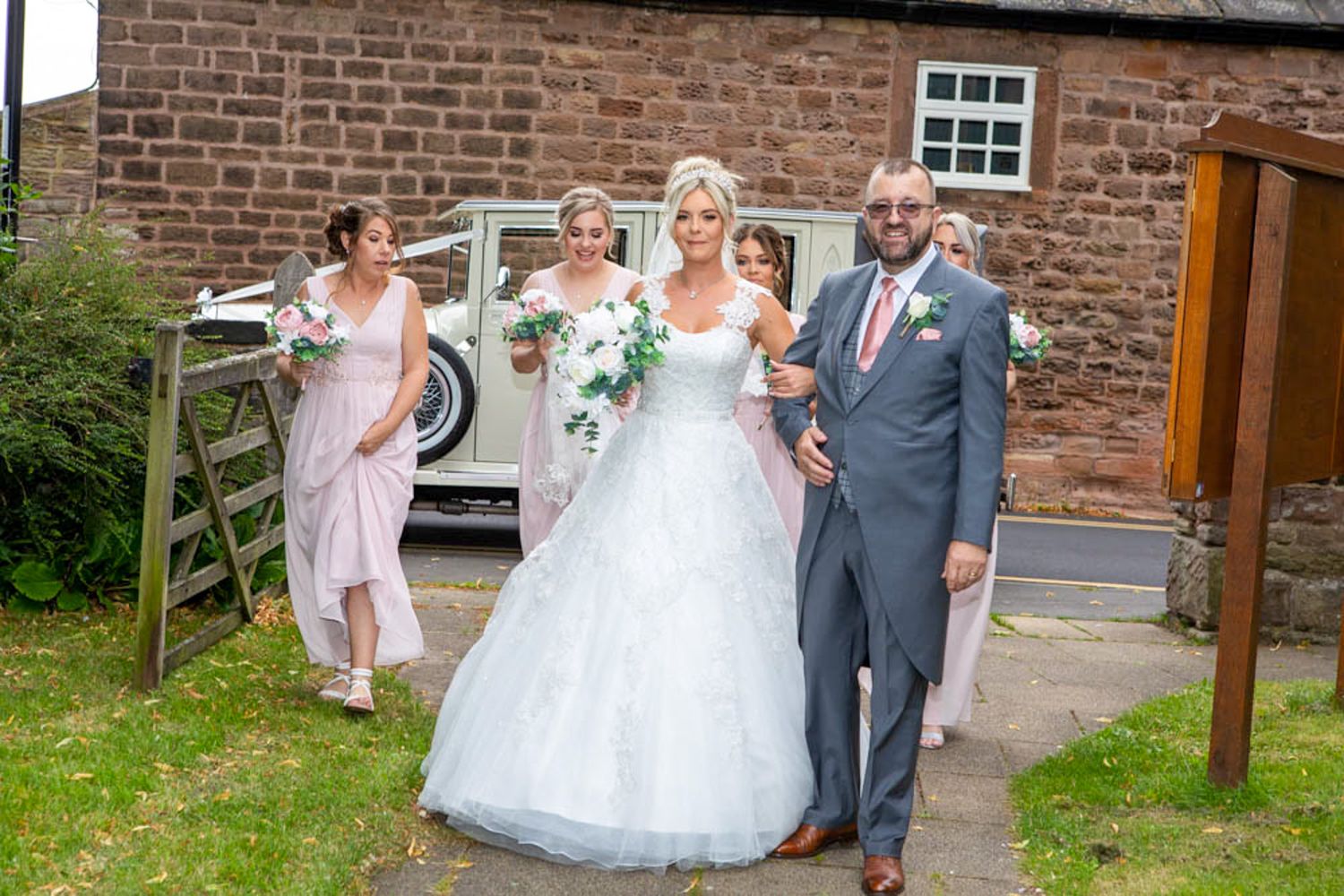 Bride in white gown with groom in gray suit and bridesmaids in pink outside rustic barn wedding venue.