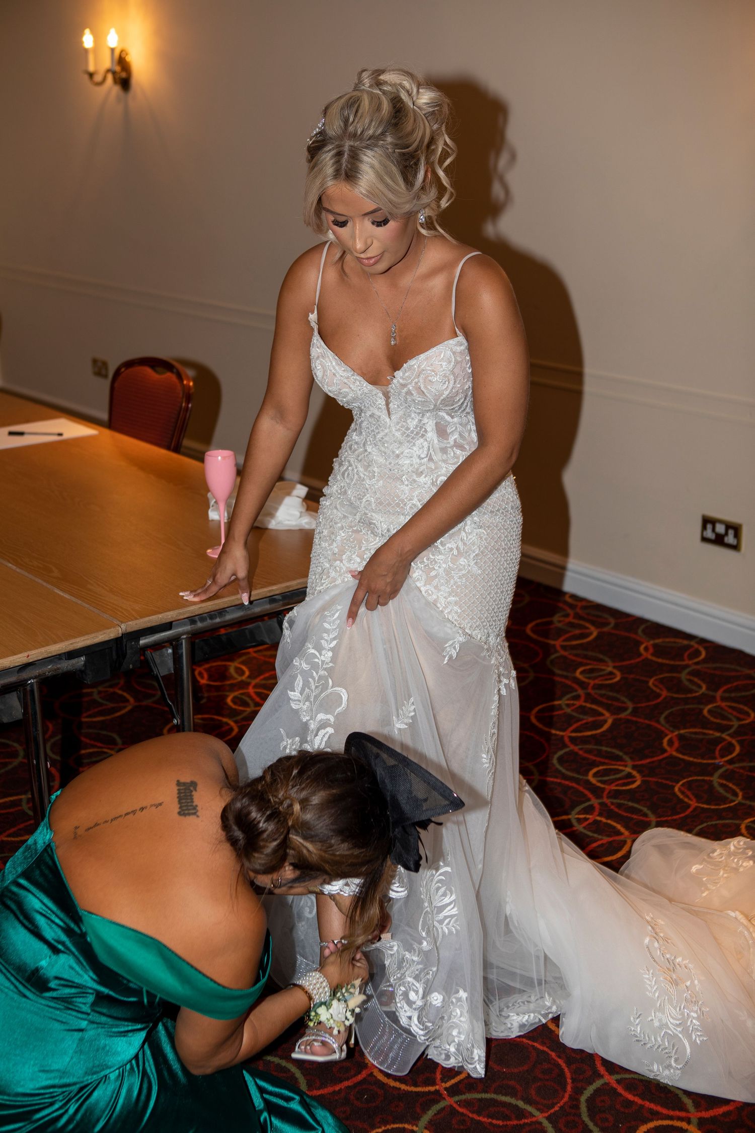 Bridesmaid in green dress adjusting bride's floral heels as she stands in elegant lace wedding gown indoors.