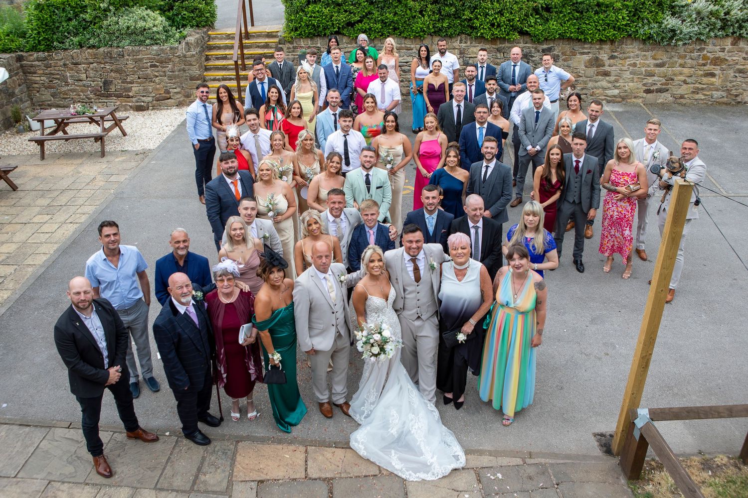 Large wedding group photo outdoors, bride and groom centered among smiling guests on a stone patio venue.