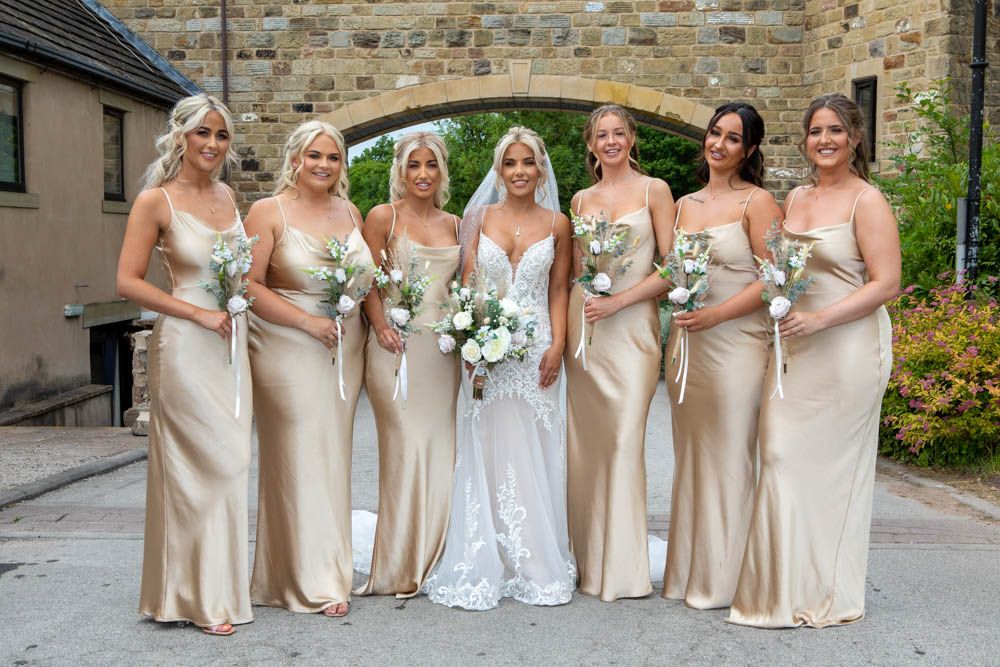Bride with six bridesmaids in gold satin dresses holding white bouquets outdoors.