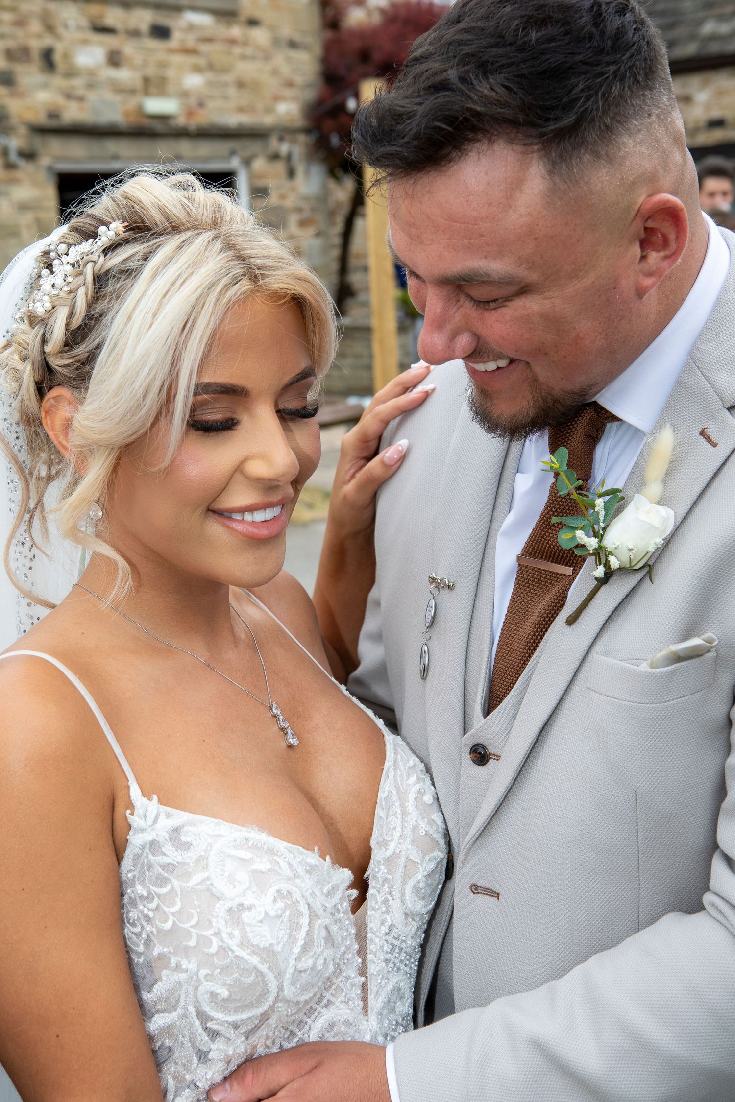A smiling bride in a white lace gown and groom in a grey suit share a tender moment at their outdoor wedding.