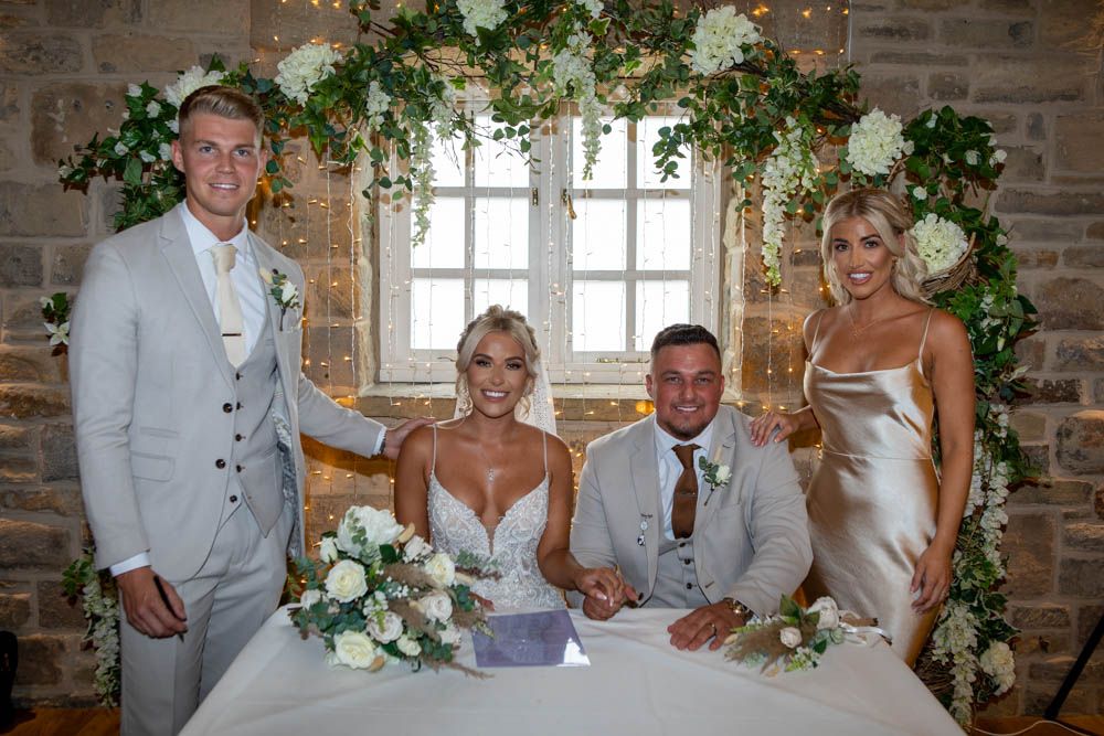 Bride and groom sign wedding register with two witnesses at floral decorated ceremony table.
