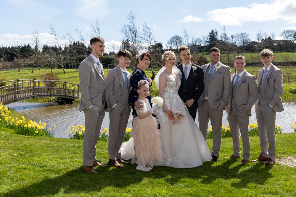 Wedding party poses outdoors on green lawn near bridge and daffodils, featuring bride, groom, groomsmen in gray suits, and flower girl.