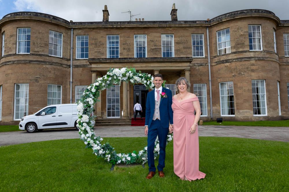 Stephen and Mich pose in front of Rudding House, Rudding Park Hotel, with a floral heart arch, man in navy suit and woman in pink gown.