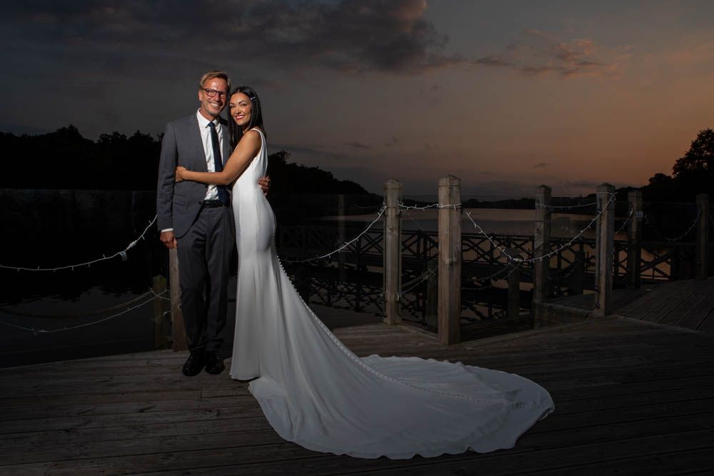 Elegant bride and groom pose on a wooden deck at sunset with a serene waterfront backdrop.
