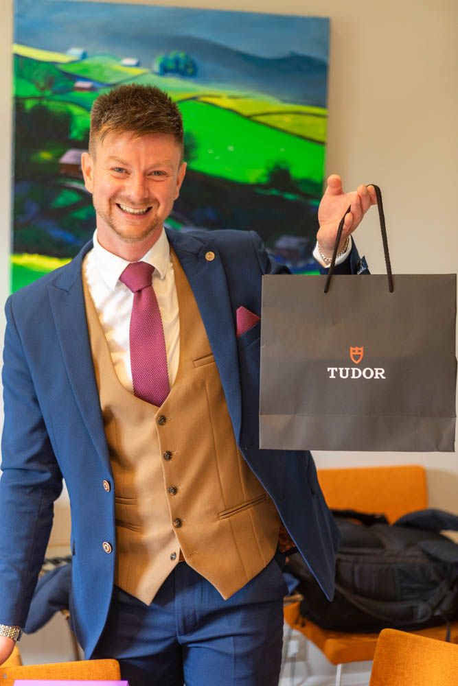 Smiling man in blue suit and tan waistcoat holding a Tudor shopping bag in front of a colorful landscape painting.