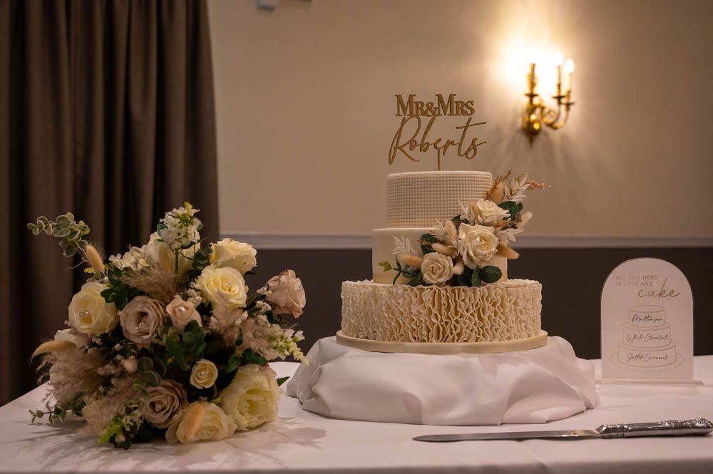 Elegant three-tier wedding cake with floral decorations and Mrs Roberts topper beside bouquet.
