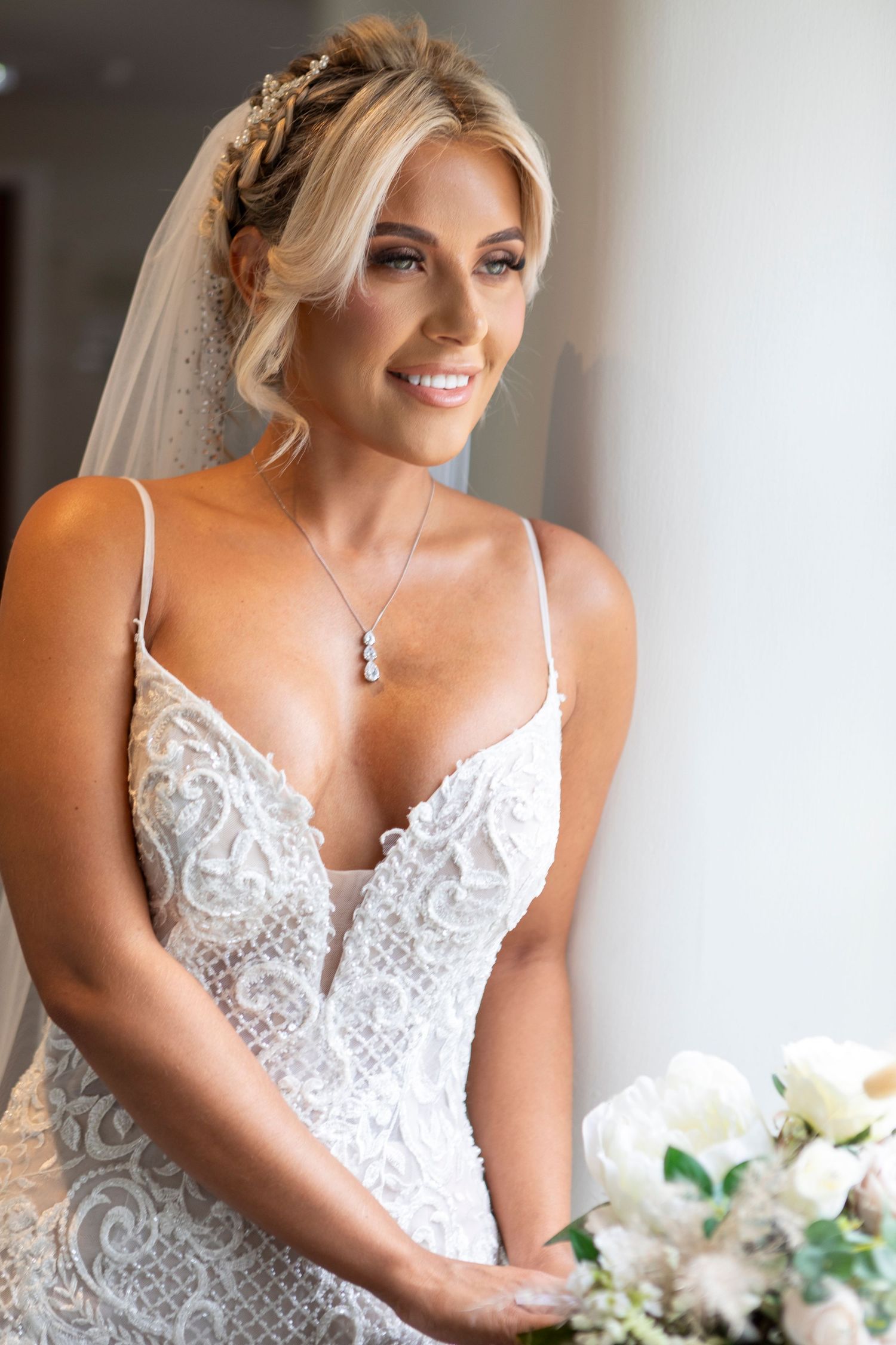 Elegant bride in lace spaghetti strap wedding gown with veil, holding white bouquet, smiling softly in natural light.