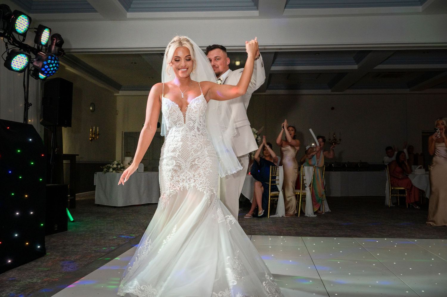 Bride in lace mermaid gown and groom in white suit share first dance at elegant wedding reception.