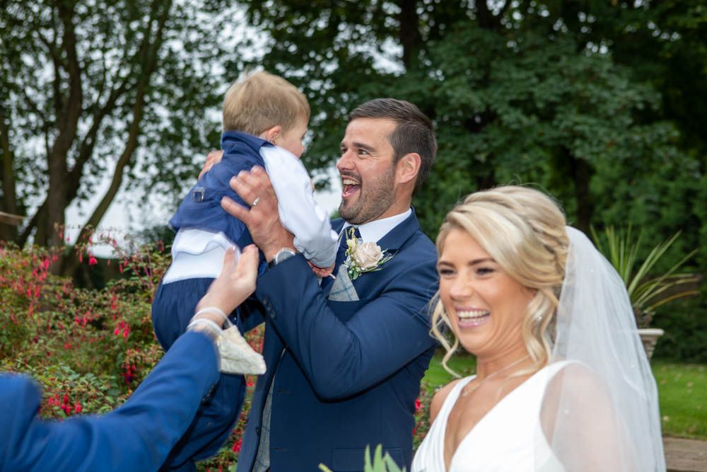 Groom in navy suit lifts laughing toddler while bride smiles in garden wedding setting.