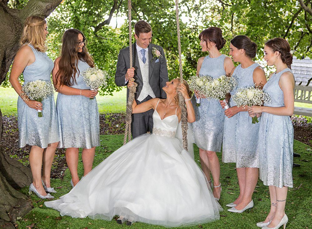 Bride on garden swing surrounded by bridesmaids in blue lace dresses holding white bouquets.