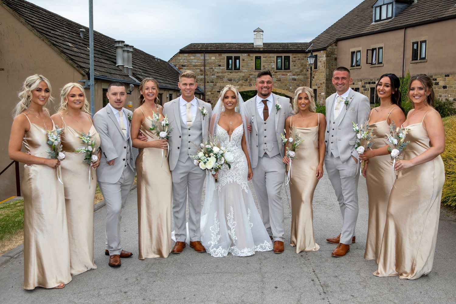 Wedding party poses outdoors, bride in white gown with groom and groomsmen in grey suits, bridesmaids in gold dresses.