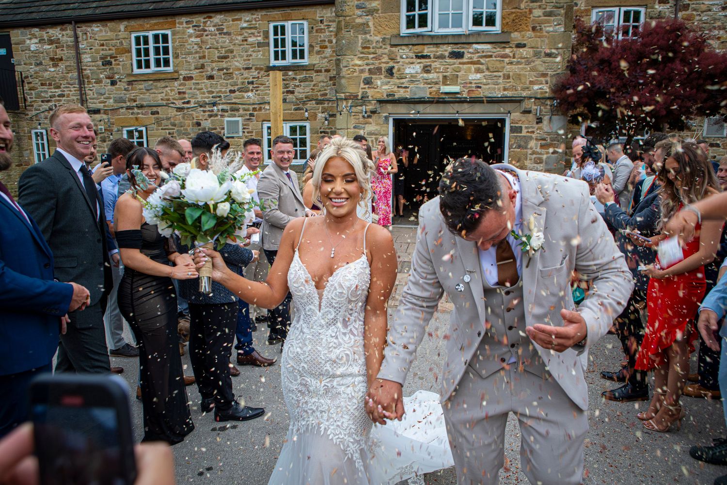 Bride and groom walk through confetti shower outside stone venue, guests celebrating joyful wedding exit.