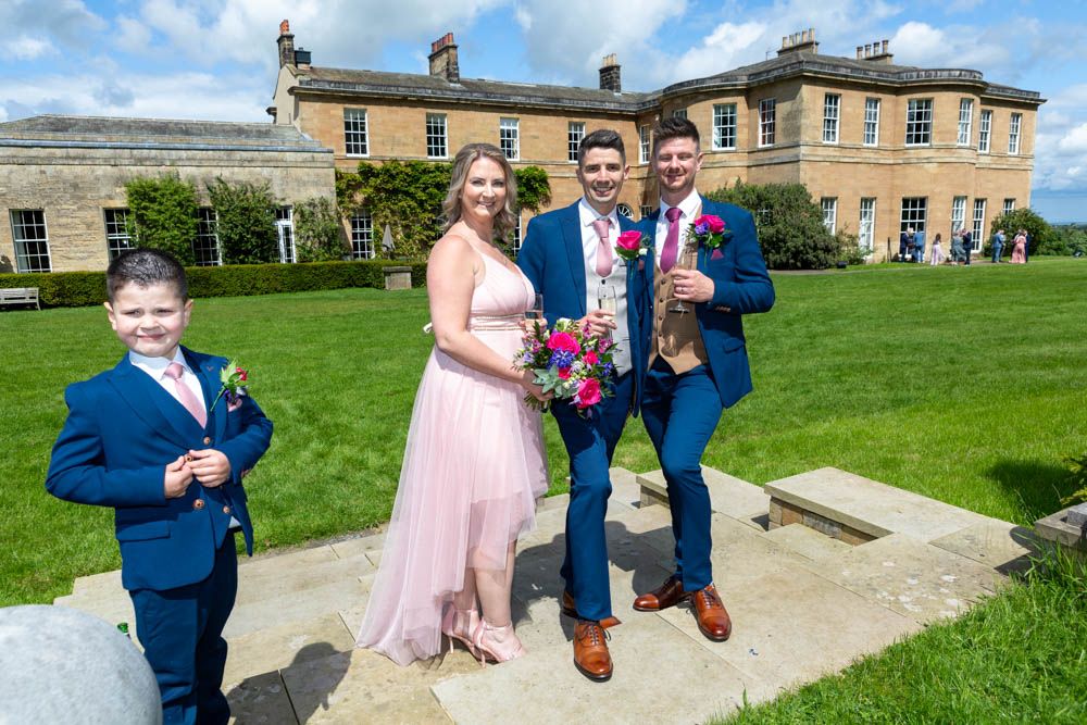Two grooms in blue suits pose with a woman in pink dress and young boy outside a Rudding House, Rudding Park Hotel.