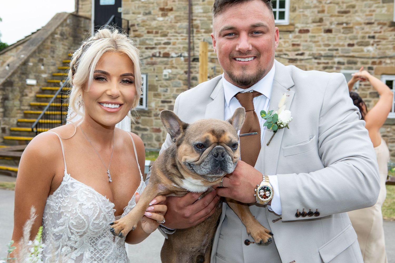 Bride and groom in wedding attire smiling and holding a French Bulldog outside a stone venue.