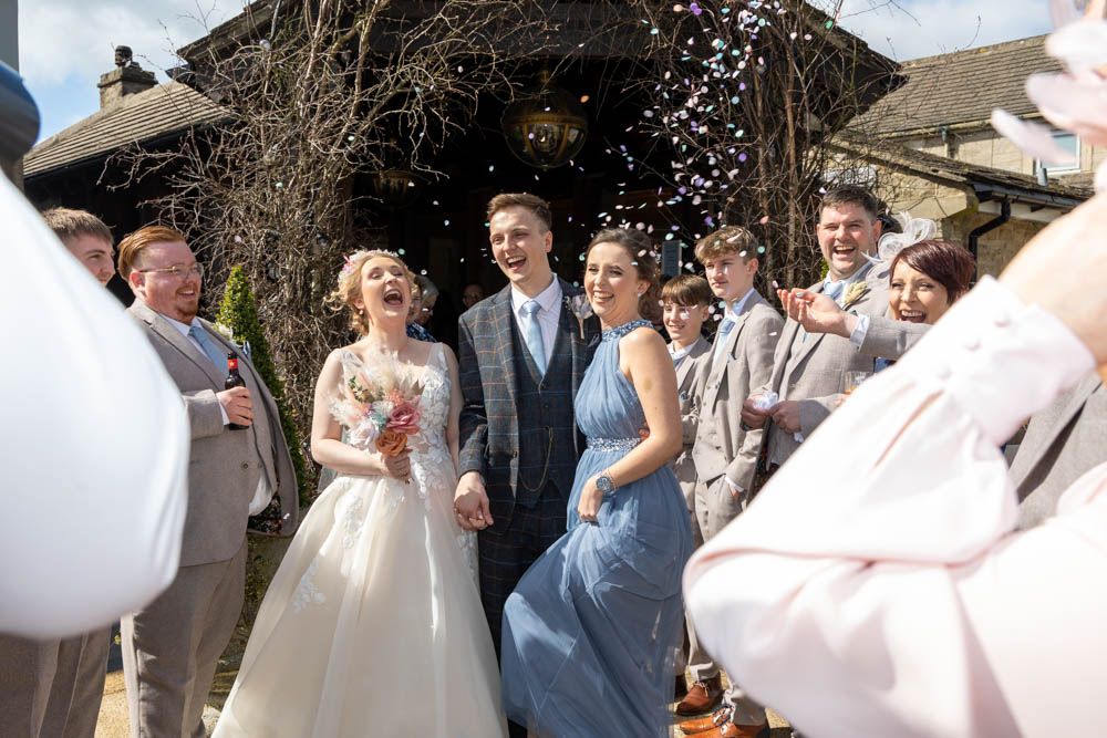 Bride and groom walk through confetti shower outside rustic stone wedding venue, surrounded by smiling guests.