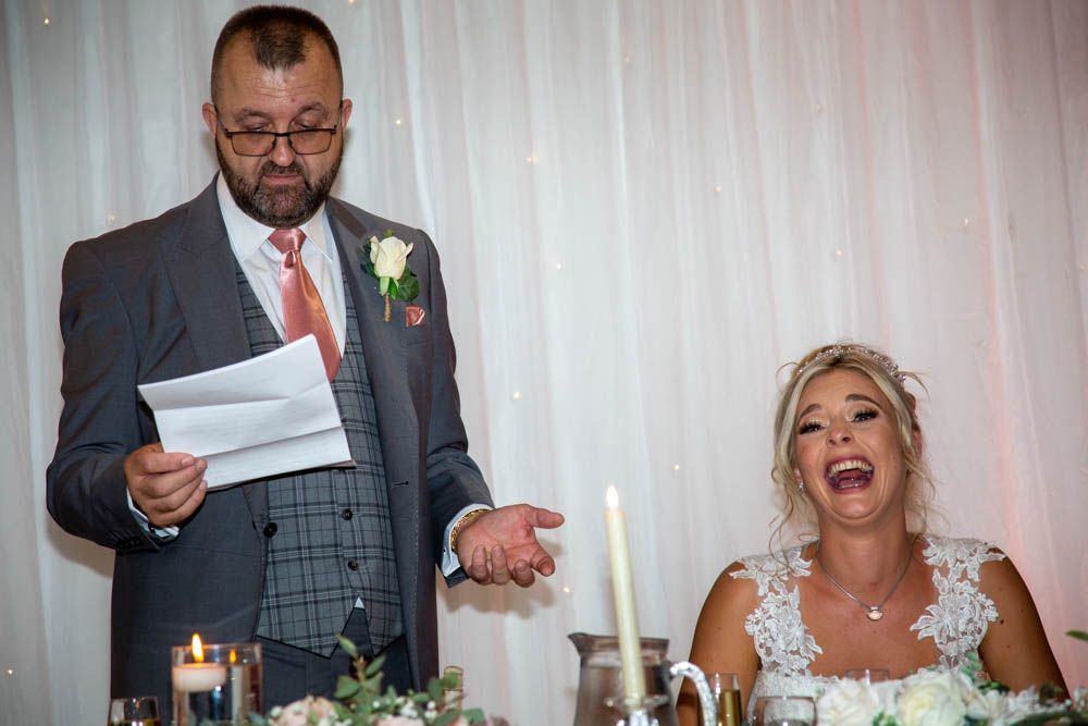 Groom reads speech at wedding reception while bride laughs joyfully, both dressed in formal attire at elegantly decorated table.