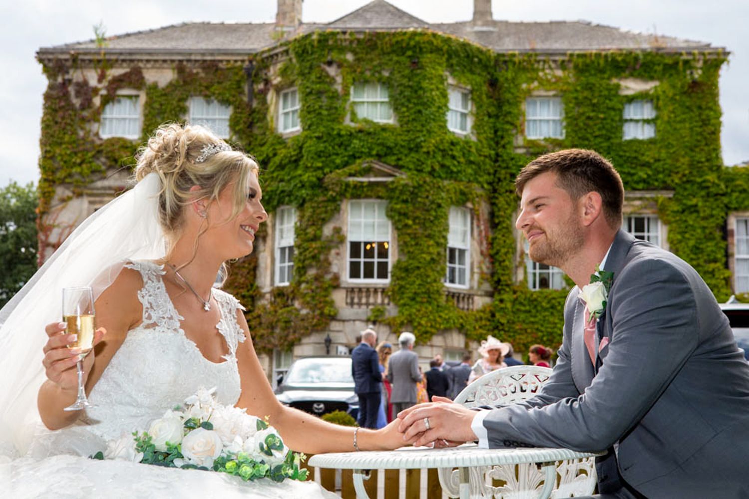 Bride in a white gown holding a champagne glass sits at a table holding hands with the groom in front of an ivy-covered manor wedding venue.