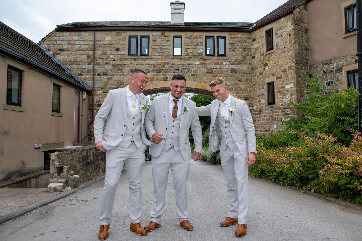 Three groomsmen in matching light grey suits and brown shoes pose outdoors at a rustic stone wedding venue.
