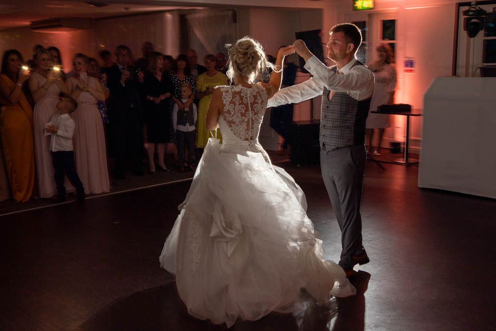 Bride and groom share their first dance at a wedding reception, guests watching in the background.