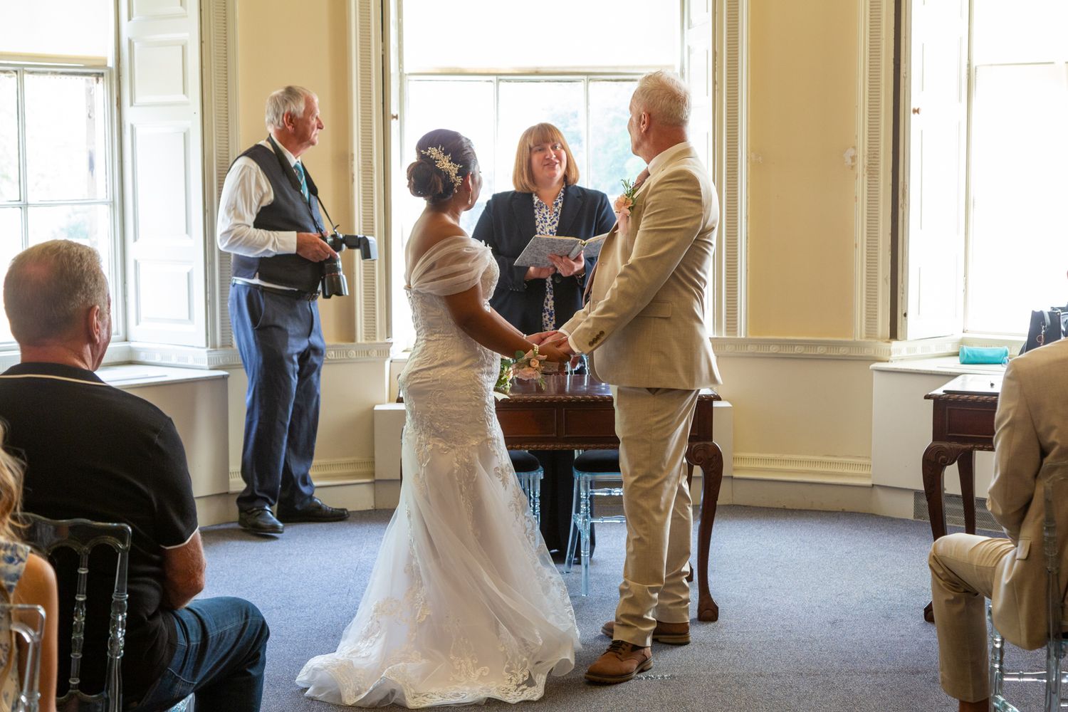 Bride and groom exchange vows before officiant in elegant room, with photographer capturing the wedding ceremony.