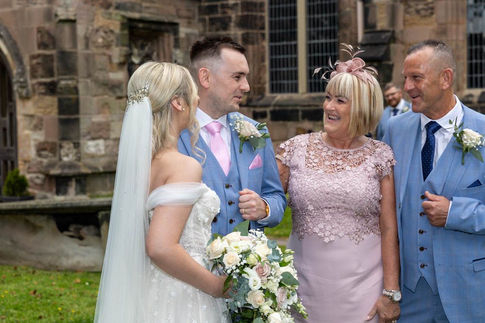 Wedding couple in white and pink attire smiling with guests outside a historic stone church.