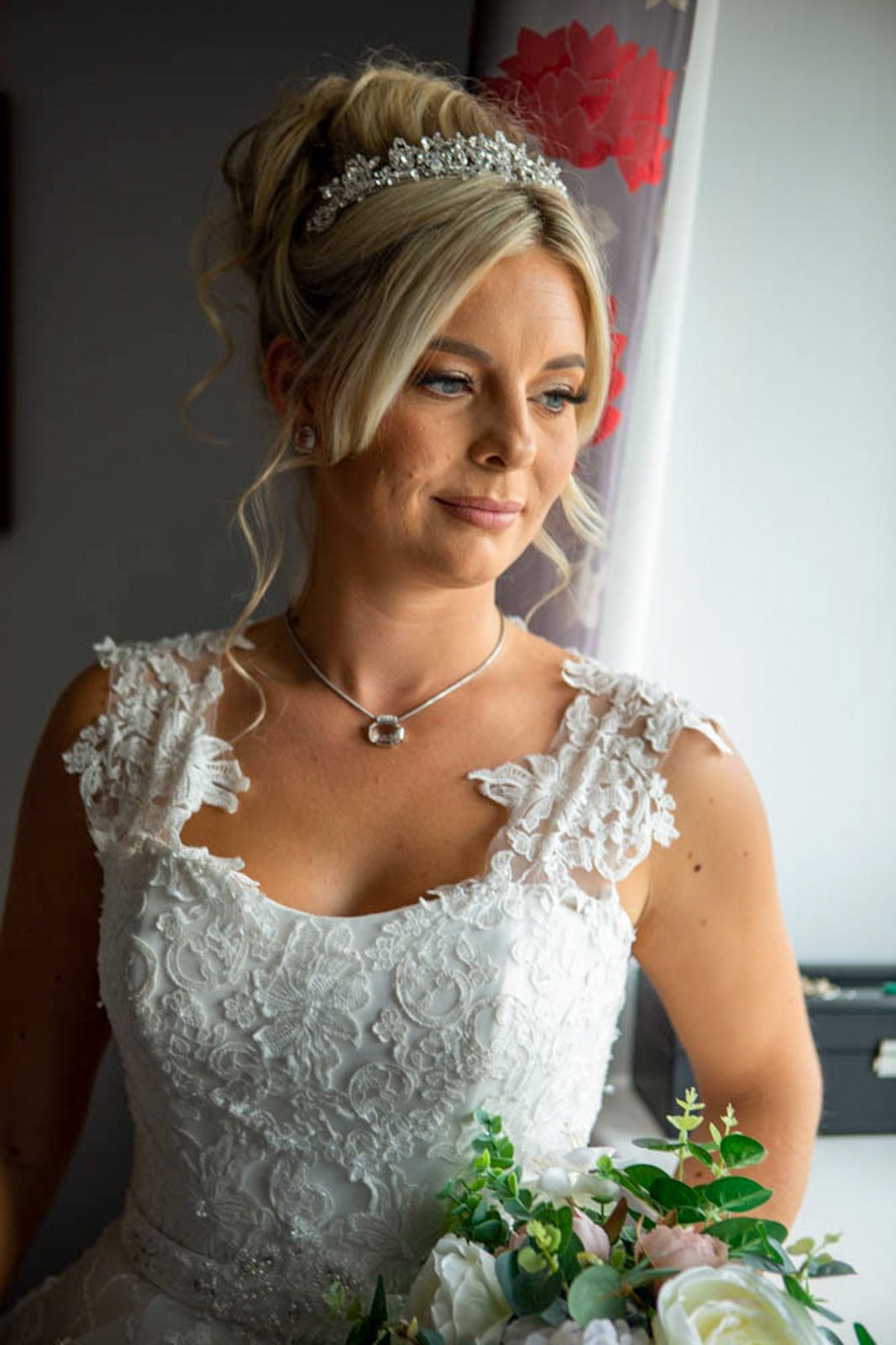 Blonde bride wearing a lace wedding dress, crystal tiara, and holding a bouquet of white and pink flowers.