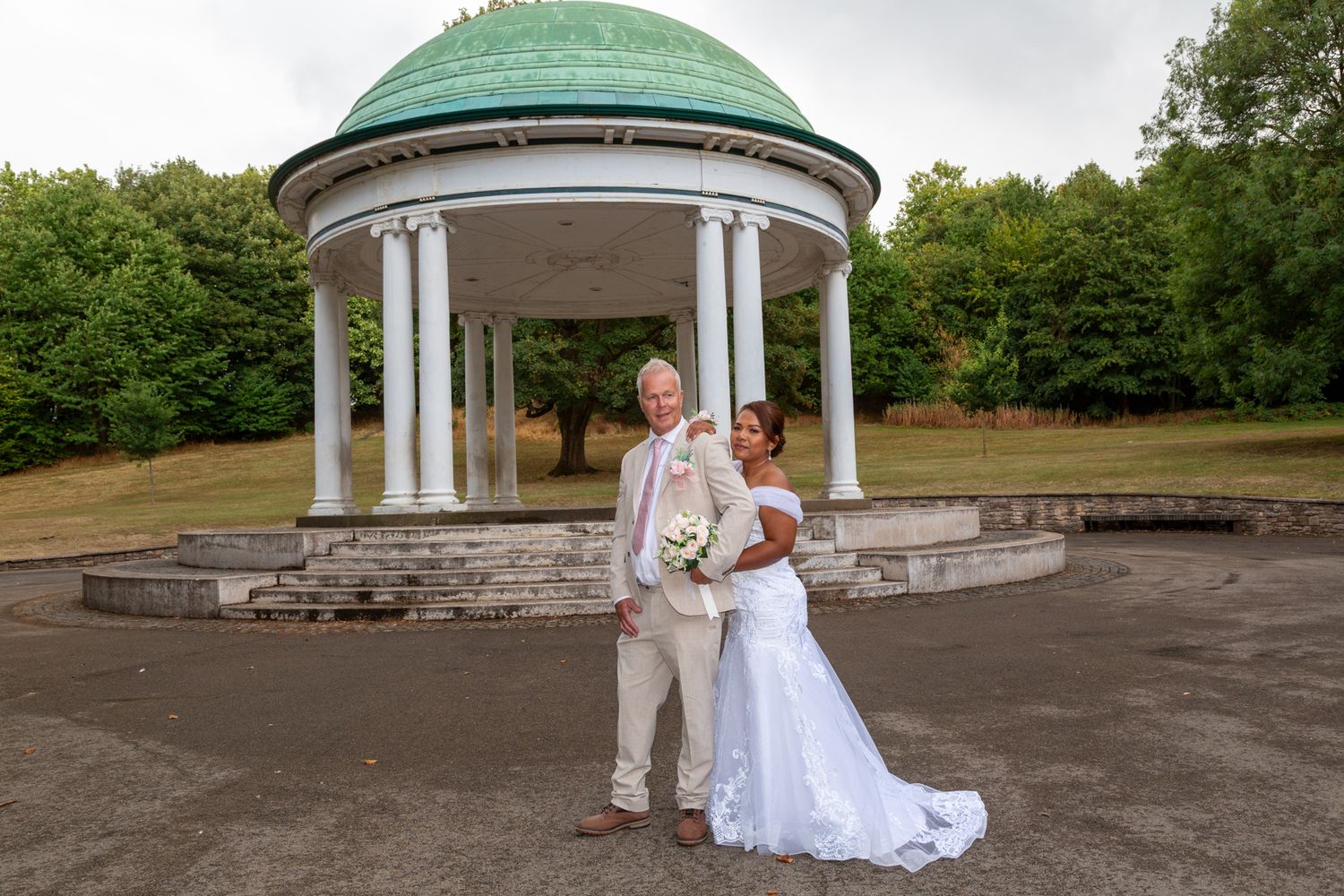 Bride and groom posing in front of a white gazebo with green dome roof surrounded by lush trees on their wedding day.