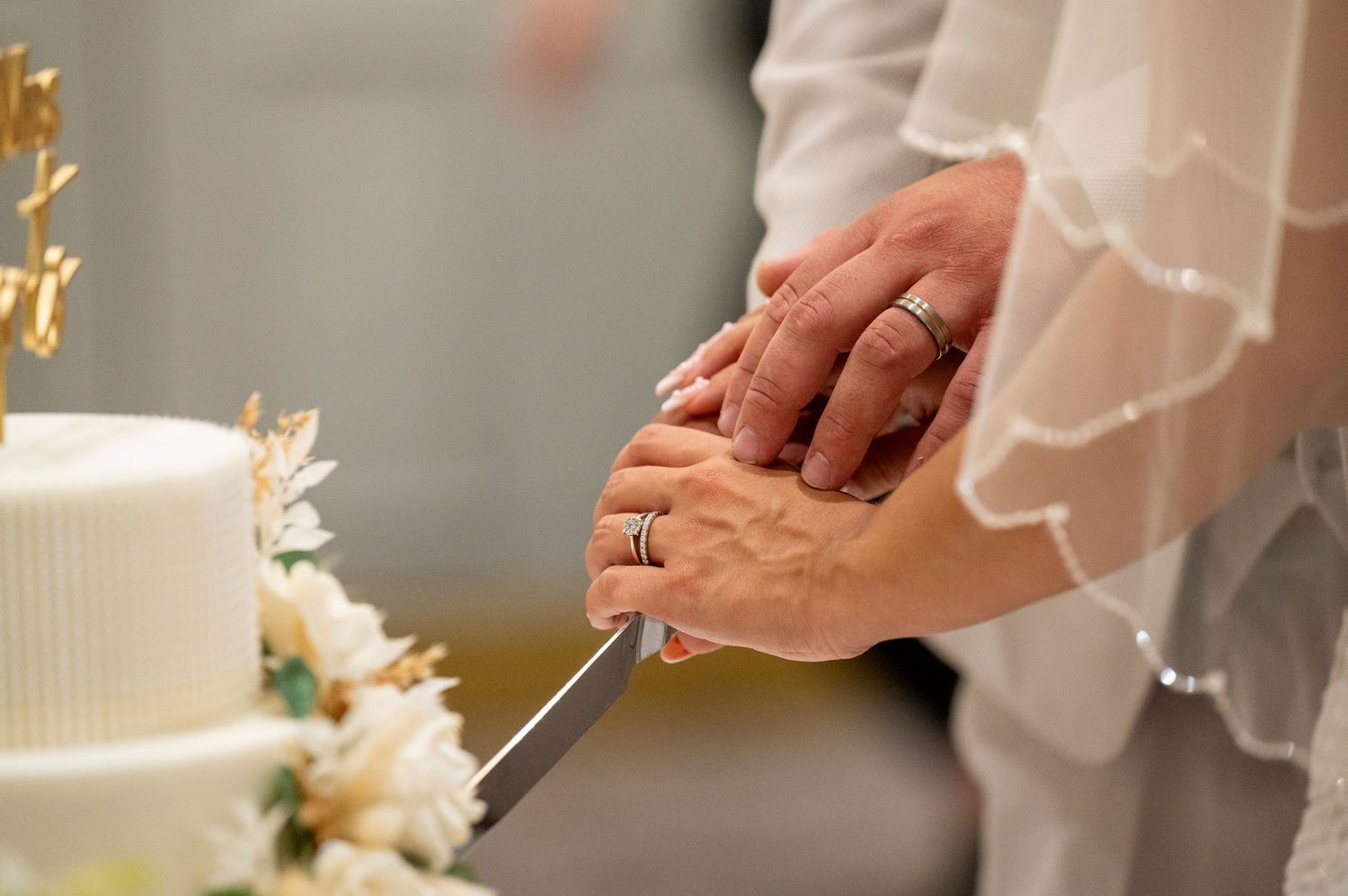 Bride and groom cutting white wedding cake together, wearing rings, with floral decorations.