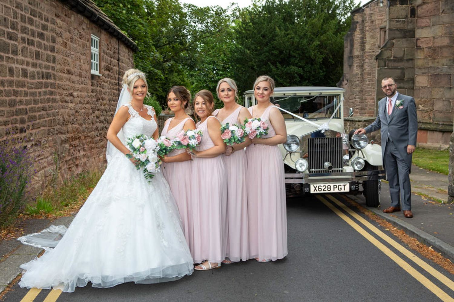 Bride with four bridesmaids in pink gowns posing by a vintage white wedding car, with groom in grey suit nearby.