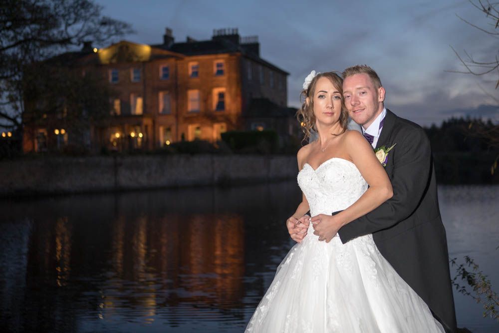 Bride and groom pose romantically by the lake at dusk with the grand Waterton Park manor house reflected in the water.