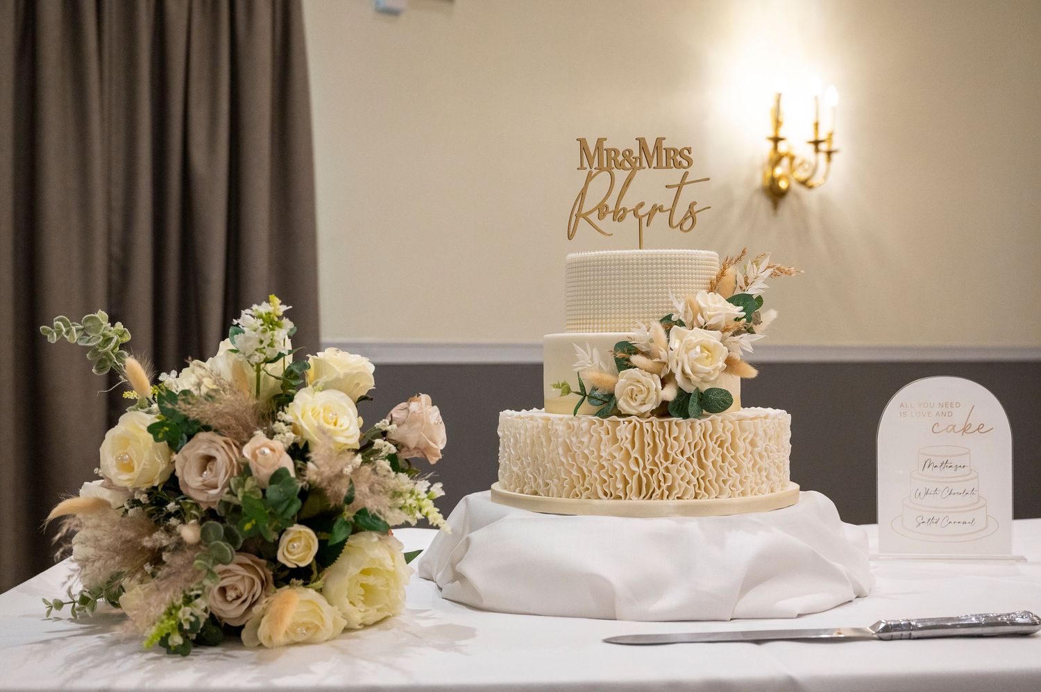 Elegant three-tier wedding cake with floral decor and Mrs & Mrs Roberts topper on a white tablecloth.