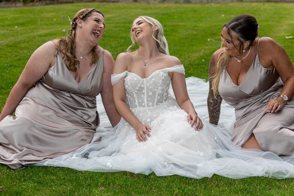 Bride in white gown laughing with two bridesmaids in silver dresses while sitting on green grass.