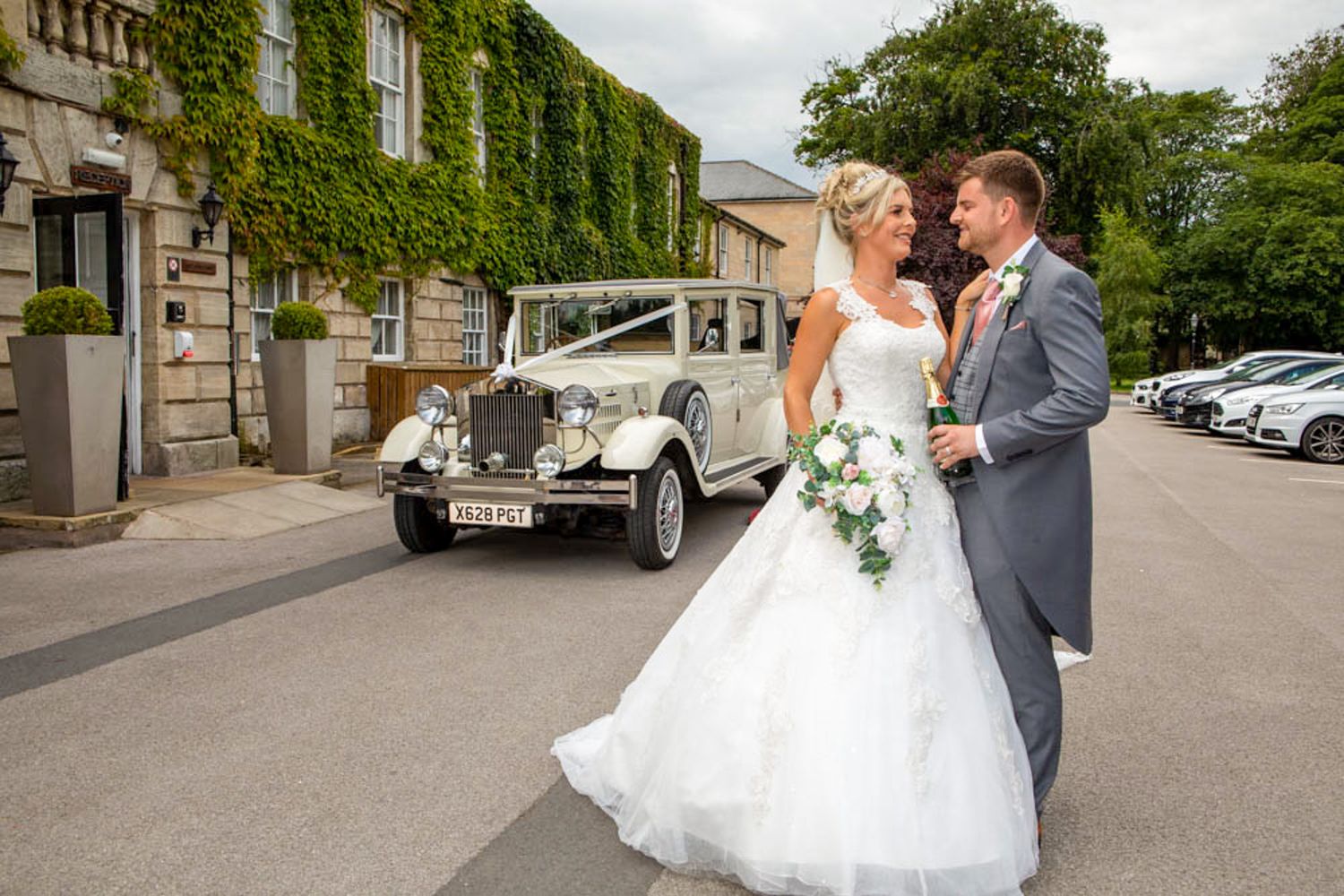 Bride and groom pose beside a vintage white wedding car outside an ivy-covered venue - Aston Hall Hotel - on their wedding day.
