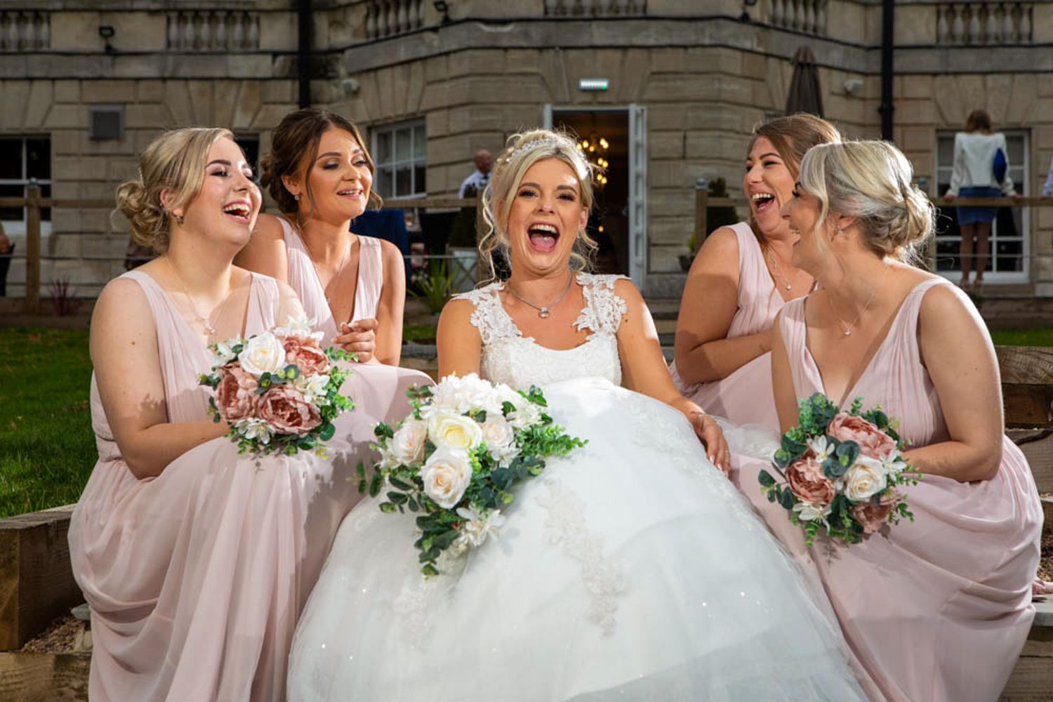 Bride in white gown laughing joyfully with four bridesmaids in blush pink dresses holding bouquets outside Aston Hall Hotel.