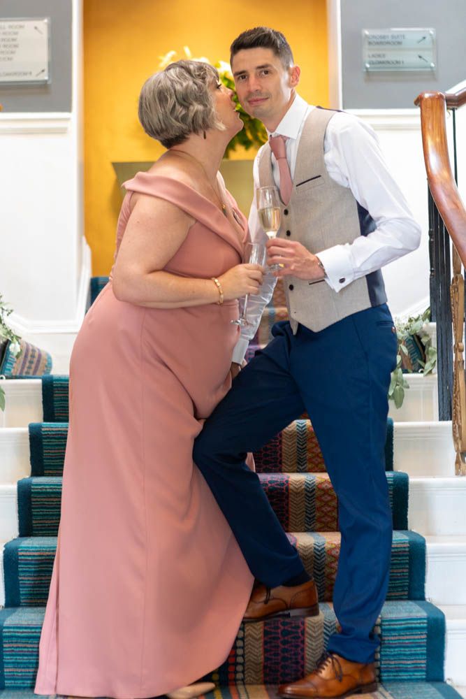 A woman in a pink gown kisses a young man in a vest on the cheek at a wedding reception on carpeted stairs.